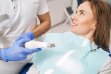 Dentist with dental device sitting next to woman in clinic