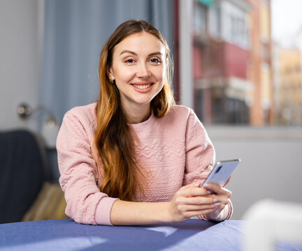 Young smiling woman sitting at a table in an apartment is texting with someone on a mobile phone