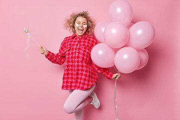 Obraz na płótnie Canvas Horizontal shot of overjoyed happy woman holds bunch of inflated balloons and magic wand exclaims gladfully feels very glad celebrates special occasion isolated over pink background. Party time