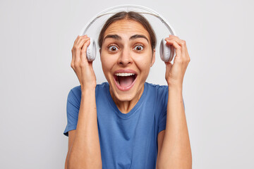 Emotional dark haired woman takes off headphones listens music with loud sound exclaims and keeps mouth widely opened stares bugged eyes dressed in casual blue t shirt isolated over white background