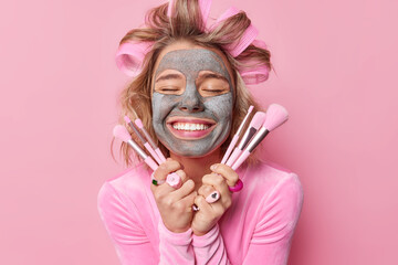 Pleased lady with hair rollers for making hairstyle smiles gently applies facial clay mask holds cosmetic brushes undergoes beauty procedures isolated over pink background. Wellness and skin care