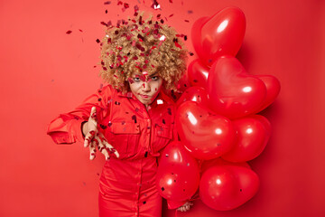 Fototapeta premium Annoyed woman doesnt like surprise received on birthday wears dress holds bunch of heart balloons angry with falling confetti on her has irritated face expression isolated over red background