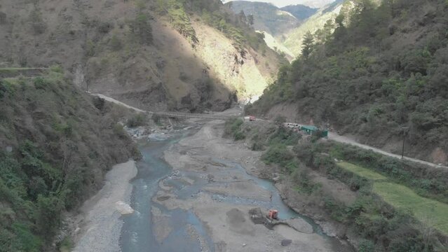 a rocky river bed excavator pulling river rock for cement production bridge spanning over water in valley connecting communities in Kabayan Benguet Philippines wide aerial approach
