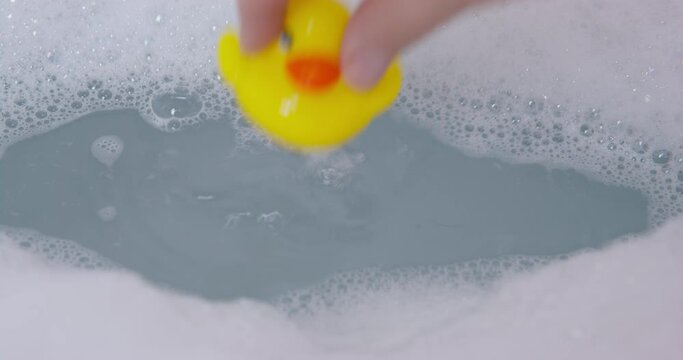 Person filled a tub with hot water and with soap bubbles, preparing it to take a relaxing bath or to bathe a child, so he threw yellow toy rubber duck into the water, close up