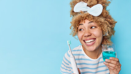 Happy curly haired young woman holds glass of fresh mouthwash and electric toothbrush undergoes hygiene procedures looks away isolated over blue background copy space to place your advertisement