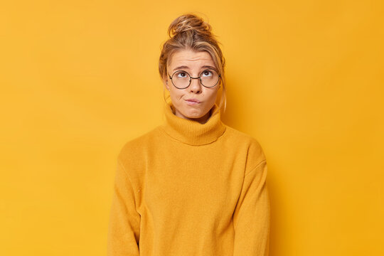 Puzzled young European bites lips focused above makes decision has bothered facial expression combed fair hair wears round eyeglasses and casual jumper isolated over vivid yellow background.
