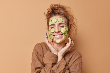 Positive young woman with combed hair applies cucumber mask on face for having clean healthy skin...