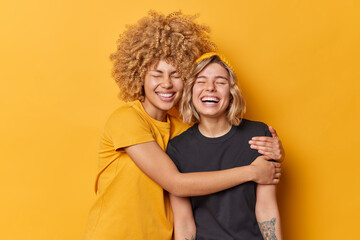 Happy female friends embrace and have good relationshipes laugh positively dressed in casual t shirts keep eyes closed isolated over yellow background. Glad sisters love each other stand indoor