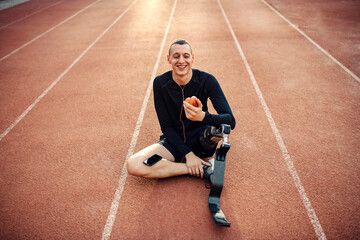 A happy handicapped runner with artificial leg taking a break, eating apple and relaxing with music.