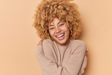 Portrait of happy young woman with curly bushy hair embraces herself feels soft dressed in casual jumper smiles broadly has white teeth isolated over beige background. Self acceptance concept