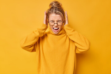 Waist up shot of emotional young blonde woman screams loudly covers ears with hands doesnt want to hear bothering sound wears spectacles and sweater isolated over yellow background. Stop this