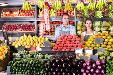 Young man and woman sellers in aprons standing near counter with the fresh vegetables and fruits on market