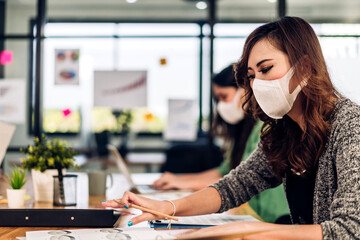 Asian businesswoman using laptop computer working and planning meeting in quarantine for coronavirus wearing protective mask with social distancing while sitting on office desk