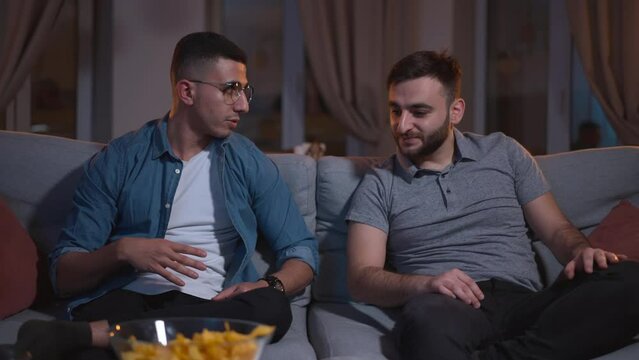 Two young men discussing match and sitting on couch in apartment interior spbas. 4k video Front view of American guys fans talk with smiles and watch football, sit on sofa in dark living room, tasty