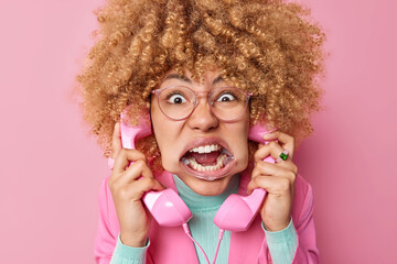 Crazy woman with curly hair holds two retro handsets near ears has telephone conversation feels annoyed keeps mouth opened shouts loudly wears transparent glasses poses against pink background