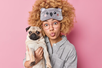 Surprised young woman with curly bushy hair keeps lips folded carries pedigree pug dog spends free...