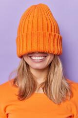 Vertical shot of positive faceless unknown woman covers eyes with knitted hat smiles broadly shows white teeth dressed casually isolated over purple background doesnt show face foolishes around