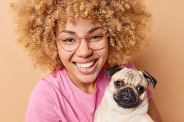 Close up shot of happy young woman with curly hair smiles gladfully enjoys company of pet poses with pug dog dressed casually isolated over beige background. People and domestic animals concept