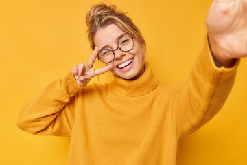 Glad young woman makes peace gesture over eye smiles broadly takes selfie wears round spectacles...