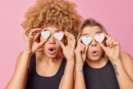 Two surprised women cover eyes with heart shaped soap keep mouthes widely opened from wonder take care of skin and personal hygiene dressed in black t shirts stand next to each other indoor.