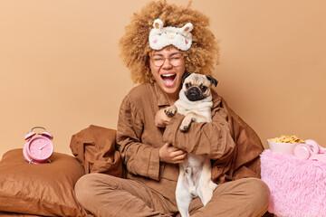 Joyful female pet owner poses in comfortable bed together with pug dog play and prepare for sleep pose in bedroom with alarmclock pillow blanket and bowl of cornflakes around. Domestic animals concept