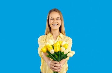 Happy young Caucasian woman isolated on solid blue background holding beautiful flowers on March 8th. Studio shot of smiling daughter or granddaughter with bouquet of yellow and white tulips in hands