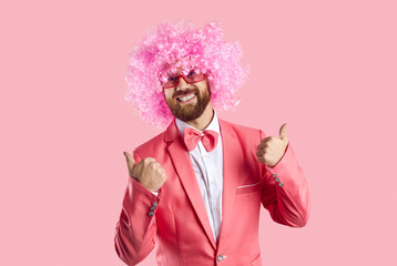 Portrait of overjoyed young man in suit and funny wig isolated on pink studio background show...