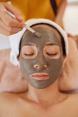 Relaxed young woman getting beauty treatment in spa salon, cosmetologist applying clay mask