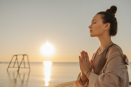 Close-up of a young fair-skinned girl doing yoga by sea during beautiful sunrise. She sits in lotus position and meditates. Rest and recovery concept.