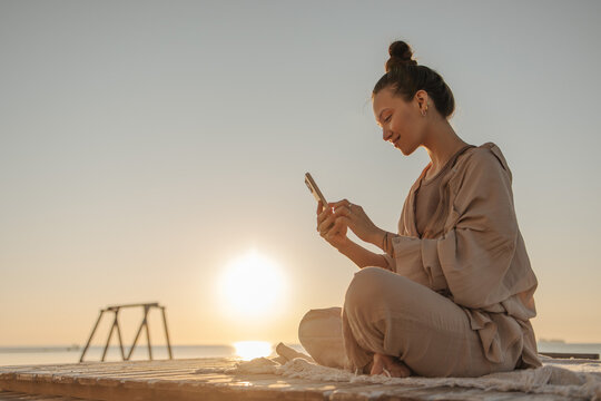 Profile photo of pretty cheerful young woman sitting cross-legged with phone against the sky. Caucasian lady with bun of hair communicates in hundreds of networks with friends, dressed casual clothes.