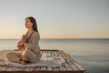 Calm young caucasian woman is meditating while sitting on wooden pier by sea. Brown-haired woman...
