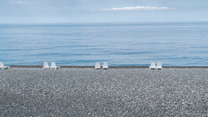 Fototapeta na wymiar Empty pebble beach with white sun loungers on a sunny day. Seascape in good weather.