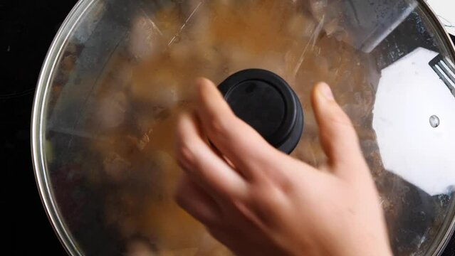 A woman's hand with a wooden spatula stirs the chicken offal being cooked in the wok and covers it with the lid. Close-up. Asian-style cooking
