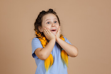 Surprised, shocked girl touching face with hands looking up with shining eyes with yellow kanekalon braids on head on beige background wearing blue t-shirt and jeans.