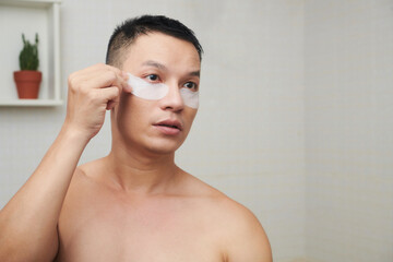 Shirtless Asian young man standing in bathroom and applying brightening and firming undereye patches
