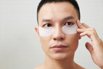 Face of serious young Asian man applying collagen under eye patches