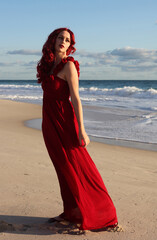 Fototapeta na wymiar Full length portrait of redhead woman wearing elegant gown. Standing pose with gestural hands at sunset ocean beach landscape background.