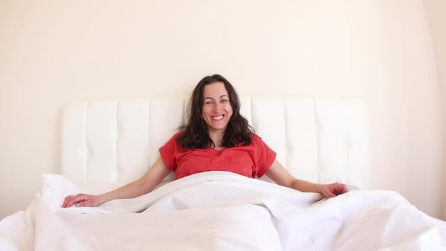 A girl in red pajamas sits on a white bed, morning awakening, portrait of a smiling woman