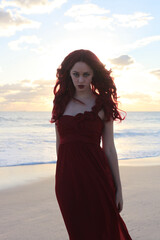 Full length portrait of  redhead woman wearing elegant gown. Standing  pose with gestural hands at sunset ocean beach landscape background.