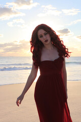 Full length portrait of  redhead woman wearing elegant gown. Standing  pose with gestural hands at sunset ocean beach landscape background.
