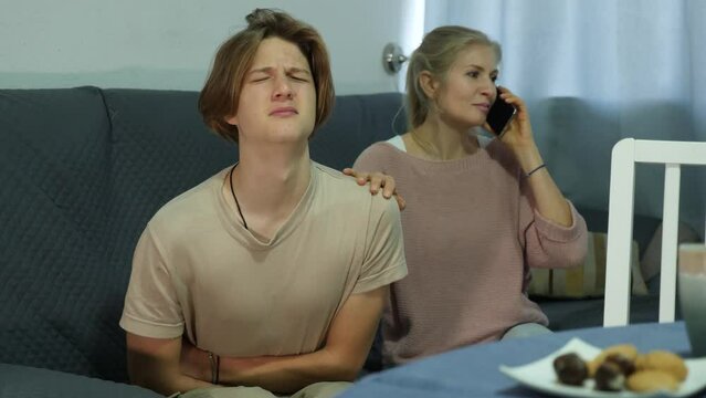 Teenage boy suffering from abdominal pain sitting on sofa at home, holding his stomach and curving through spasms while concerned mother speaking on phone with doctor
