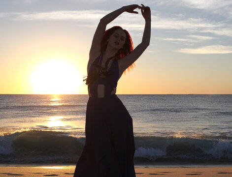 Full length portrait of  redhead woman wearing elegant gown. Standing  pose with gestural hands at sunset ocean beach landscape background.