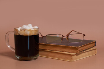 A glass cup of coffee with marshmallows, stack of old books and glasses