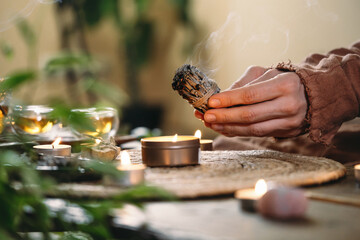 Woman hands burning white sage, before ritual on the table with candles and green plants. Smoke of...