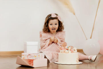 Fototapeta na wymiar A cute little girl is sitting on the floor with a cake and gifts on her birthday and eating a lollipop