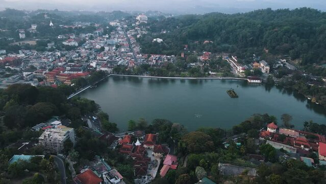 A large group of white birds flies low over Kandy Lake in Sri Lanka between the hilly landscape with extensive nature and the densely built village in the background. Backwards drone dolly shot