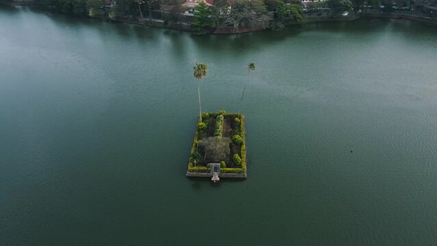Beautiful square Diyathilaka Mandapaya with nice gardens and two tall thin palm trees in the reflecting Kandy Lake in Srilanka as multiple birds fly around. High angle drone panning shot