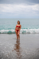 A middle-aged woman with a good figure in a red swimsuit on a pebble beach, running along the shore in the foam of the waves