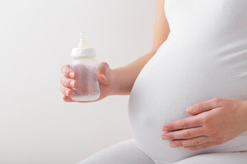 Young adult pregnant woman hand holding plastic bottle for milk on light gray background. Preparing feeding things for future newborn in pregnancy time. Closeup. Baby expectation. - obrazy, fototapety, plakaty