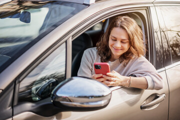 Cheerful young woman sitting in a car in the driver's seat looking into a smartphone, paying for...